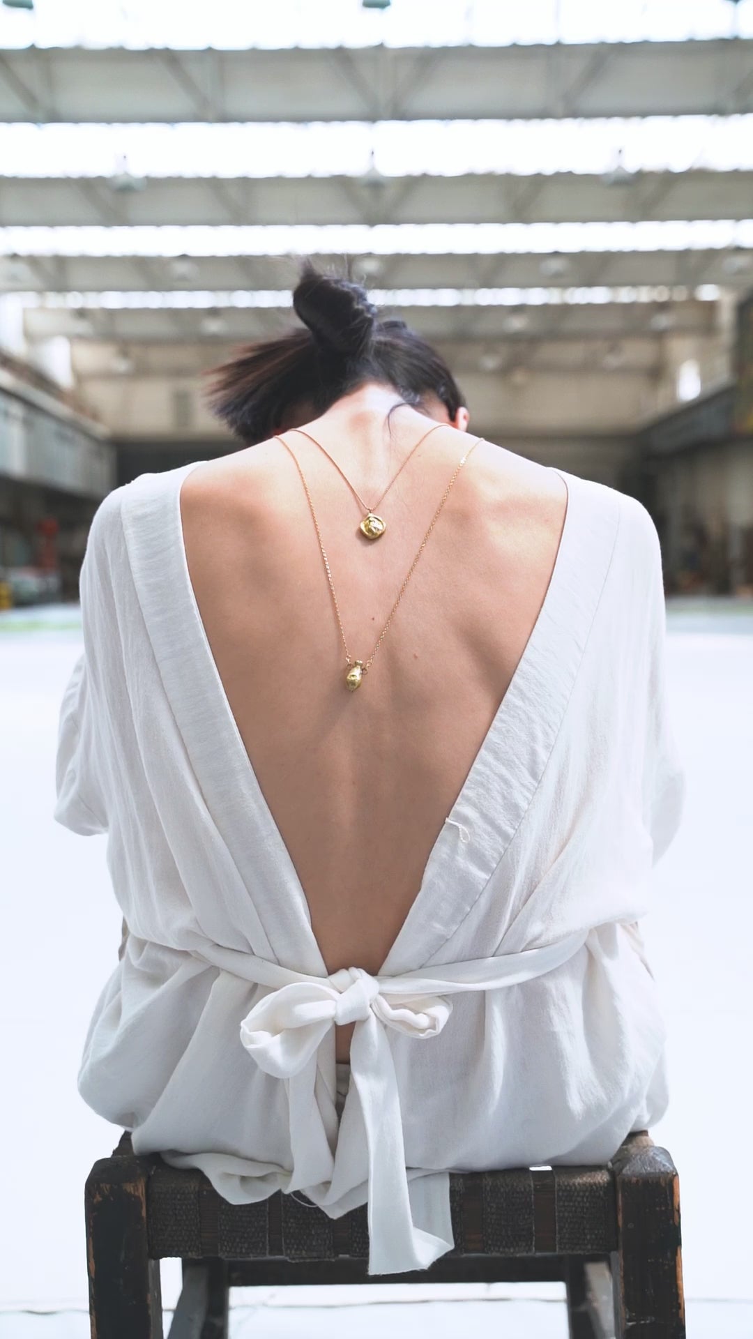 a video showcasing jewelry on a model sitting on a chair with her back to the camera wearing two necklaces with pendants on her back